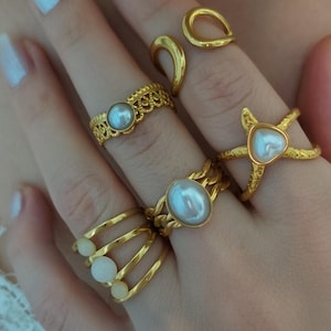 Op de afbeelding: Een hand met meerdere gouden ringen. De ringen hebben verschillende ontwerpen, waaronder parels en ingewikkelde patronen. Stijlen zijn onder meer een ring in de vorm van een zeester en een ring met drie banden met kleine witte stenen.