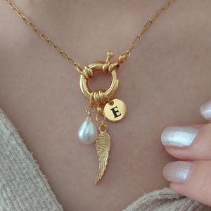Op de afbeelding: Een goudkleurige ketting met een dikke schakelketting en een ronde sluiting. Aan de sluiting hangen drie bedels: een witte druppelvormige parel, een gestructureerde goudkleurige vleugel en een kleine ronde goudkleurige schijf met de letter E.