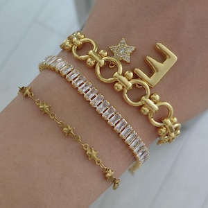 Op de afbeelding: Een set van drie goudkleurige armbanden. Eén heeft een fijne ketting met kleine sterbedels, een andere is een tennisarmband met rechthoekige doorzichtige stenen, en de derde is een schakelarmband met een sterbedel en de letter E.