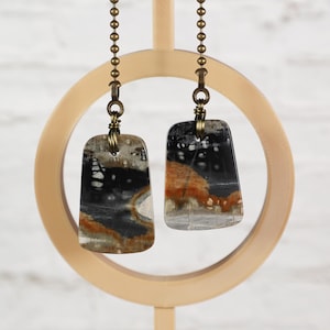 May include: Two rectangular pendants made of polished stone with natural patterns in shades of black, white, and rust orange. Each pendant is suspended from a bronze-toned metal ball chain by a wire-wrapped loop at the top of the stone piece.