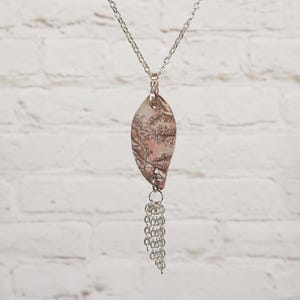 May include: A silver chain necklace featuring a leaf-shaped pendant with a muted pink and brown botanical print. The pendant is accented with a dangling silver chain tassel at the bottom, creating a layered look that hangs from a delicate metal link chain.