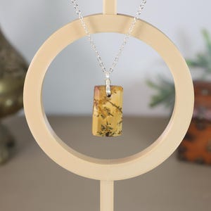 May include: A silver chain necklace with a rectangular pendant. The pendant is a mix of yellow, brown, and black colors. The necklace is displayed on a cream-colored circular stand, with a blurred background.