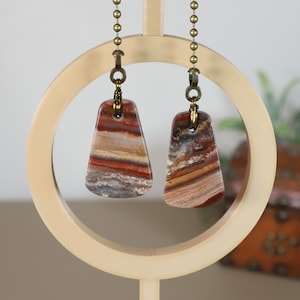 May include: Two rectangular pendant earrings with a layered, earthy color scheme of red, brown, and cream. The pendants hang from brass-colored chains and are displayed on a light-colored wooden stand. The earrings have a natural, stone-like appearance.