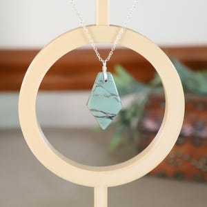 May include: A turquoise-colored pendant necklace with a silver chain. The pendant is a geometric shape with dark veining. The necklace is displayed on a cream-colored circular stand, with a blurred background of greenery and a wooden cabinet.