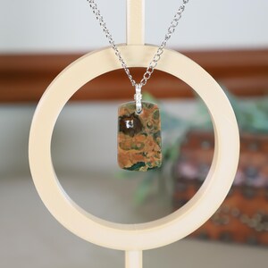 May include: A rectangular pendant necklace with a silver chain. The pendant is a mix of brown, orange, and green tones. The necklace is displayed on a cream-colored stand with a circular design. The background is blurred, with hints of brown and green.
