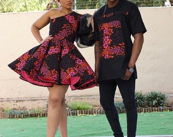 May include: A couple wearing matching black and pink patterned outfits. The woman is wearing a short, one-shoulder dress with a pink underskirt and a matching head wrap. The man is wearing a long-sleeved shirt with a large letter "L" on the front.