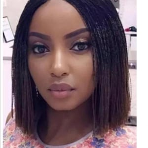May include: A black wig with small braids, styled in a bob haircut. The wig is worn by a person with dark skin and features a middle parting.