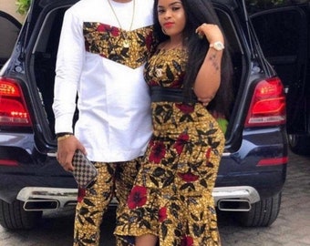 May include: A couple wearing matching outfits with a floral print. The man is wearing a white shirt with a floral print panel and matching pants. The woman is wearing a floral print dress with a black belt. They are standing in front of a black car.