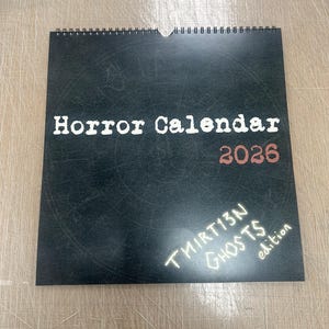 May include: A black spiral-bound calendar with the words "Horror Calendar 2026" in white and red lettering. The bottom right corner features the text "THIRT13N GHOSTS edition" in white, suggesting a themed calendar.