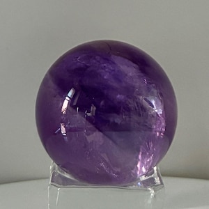 Può includere: Una sfera di ametista viola lucidata con inclusioni interne visibili e variazioni di colore naturali, che poggia su un supporto espositivo quadrato in vetro trasparente che fornisce una base stabile per la pietra rotonda.