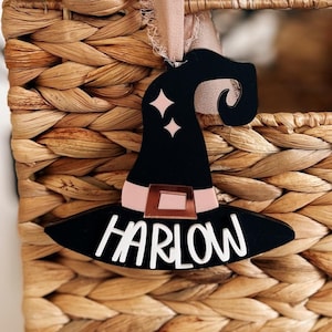 May include: Black witch hat ornament with a pink ribbon and the name "HARLOW" written in white on the brim. The ornament is hanging from a wicker basket.