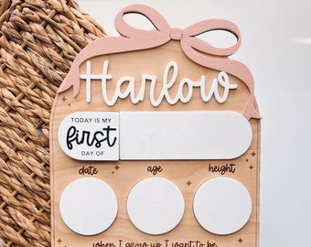 Peut inclure: Un tableau de croissance en bois avec un nœud rose et le nom "Harlow" écrit en blanc. Le tableau comporte des espaces à remplir avec la date, l'âge, la taille et ce que l'enfant veut être lorsqu'il sera grand.