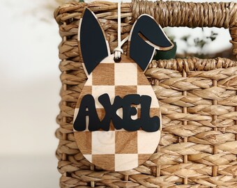 May include: An Easter-themed wooden tag in the shape of an egg with bunny ears. The tag features a checkered pattern and the name "AXEL" in black letters. The tag is hanging on a woven basket.