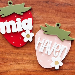 May include: Two strawberry-shaped wooden ornaments. One is red with the name "mia" in white, and the other is pink with the name "haven" in white. Both have green leaf tops and white flower accents.