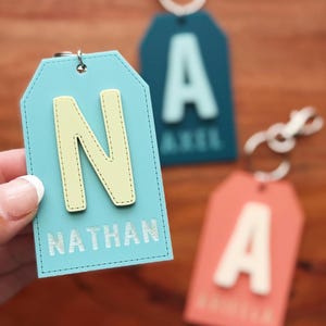 Könnte beinhalten: Nahaufnahme von drei personalisierten Gepäckanhängern. Der vordere Anhänger ist hellblau mit einem gelb genähten Buchstaben "N" und dem Namen "NATHAN" in Silber. Weitere Anhänger sind türkis und korallenfarben mit weißen Buchstaben.