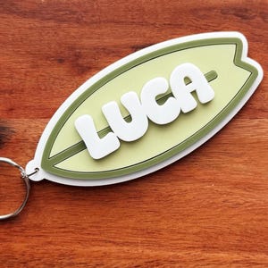 May include: A surfboard-shaped keychain with the name "LUCA" in white, raised letters. The surfboard is light green with a dark green outline and a silver keyring. The background is a wooden surface.