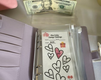May include: A light purple binder with a clear plastic sleeve containing a "Mini Dollar Savings Challenge" sheet. The sheet has heart shapes with dollar amounts. A twenty-dollar bill is visible above the binder.
