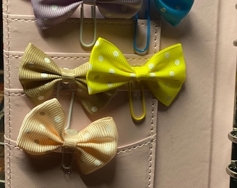 May include: A collection of colorful bow paper clips. The bows are in shades of lavender, blue, yellow, tan, and peach, each with white polka dots. The clips are attached to a pink leather notebook.