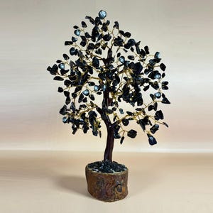 Könnte beinhalten: Eine dekorative Baumskulptur mit einem dunkelbraunen Stamm und einem Sockel aus rauem, strukturiertem Material. Die Äste des Baumes sind aus goldfarbenem Draht gefertigt, und die Blätter sind kleine, dunkelblau-schwarze Steine. Der Baum ist etwa 20 cm hoch.