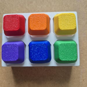 May include: A set of six square keyboard keycaps arranged in a white plastic tray. The keycaps feature a textured surface and are coloured in red, orange, yellow, purple, blue, and green. Each keycap has a smooth, rounded edge design.