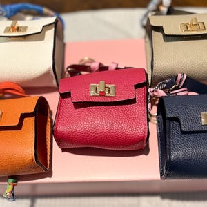 Puede incluir: Cinco pequeños bolsos cuadrados en varios colores: blanco, naranja, rojo, beige y azul marino. Cada bolso tiene una superficie texturizada, un cierre dorado y un pequeño asa o detalle de bufanda. Están dispuestos sobre una superficie rosa.