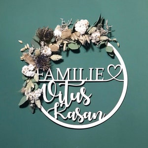 Könnte beinhalten: Weißer Holz-Kreis mit den Worten "Familie Vitus Kasan" und einem Herzsymbol, verziert mit getrockneten Blumen und Grün.