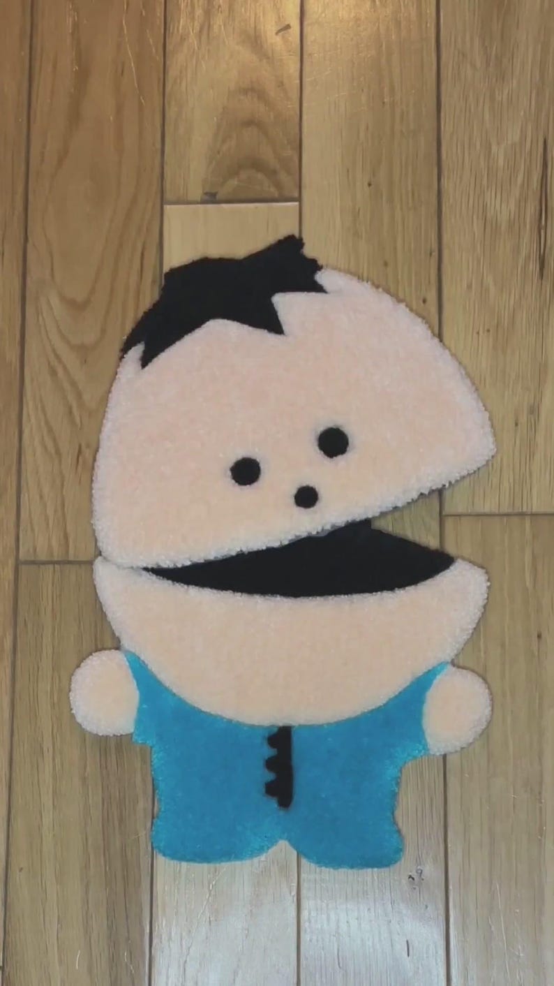 May include: A plush toy of a cartoon character with black hair, a blue shirt, and a large open mouth. The toy is made of soft, fuzzy fabric and has a light brown skin tone.