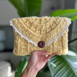 Op de afbeelding: Een gele gehaakte clutch met een witte gehaakte rand en een bruine knoopsluiting.
