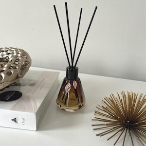 May include: A brown glass diffuser bottle with gold leaf accents and black diffuser sticks. The bottle is sitting on a white surface with a book and a gold metal decorative object.