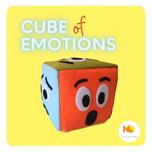 May include: A plush cube toy with different facial expressions representing emotions. The cube has blue, orange, and green sides, with the text "CUBE of EMOTIONS" above it. The toy is designed for educational play.