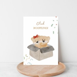 Könnte beinhalten: Eine Grußkarte mit zwei Cartoon-Teddybären in einem Karton. Ein Bär hat eine rote Schleife. Der deutsche Text "Glück IM DOPPELPACK" ist in goldener Schrift über den Bären geschrieben, mit bunten Konfetti-Akzenten.