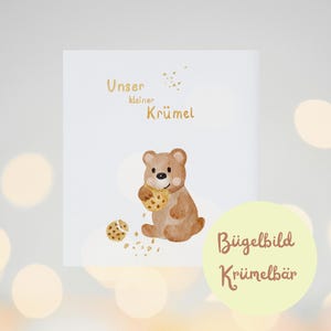 Könnte beinhalten: Ein brauner Bär mit einer hellbraunen Nase und hellbraunen Augen isst einen Keks. Der Bär sitzt auf einem weißen Hintergrund mit dem Text "Unser kleiner Krümel" und "Bügelbild Krümmelbär" in goldener Schrift.