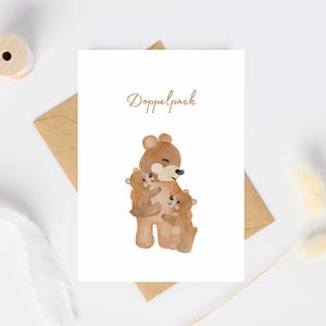 Könnte beinhalten: Aquarellillustration auf einer weißen Karte mit einem braunen Bären, der zwei kleinere Bären umarmt. Der Text "Doppelpack" ist in einer Schreibschrift über den Bären geschrieben. Die Karte liegt auf einer weißen Oberfläche mit einem braunen Umschlag.