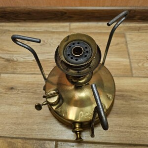 K&ouml;nnte beinhalten: Ein antiker Campingkocher aus Messing mit drei Metallbeinen und einem runden Brenner. Der Kocher hat einen schwarzen Metalldeckel mit einem kleinen Loch in der Mitte.