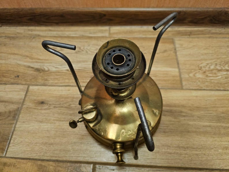 K&ouml;nnte beinhalten: Ein antiker Campingkocher aus Messing mit drei Metallbeinen und einem runden Brenner. Der Kocher hat einen schwarzen Metalldeckel mit einem kleinen Loch in der Mitte.