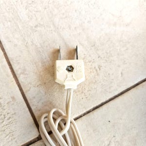 May include: A white electrical plug with two prongs and a white cord. The plug is on a tiled surface.