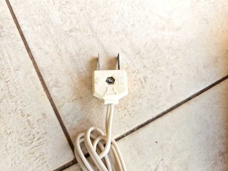May include: A white electrical plug with two prongs and a white cord. The plug is on a tiled surface.