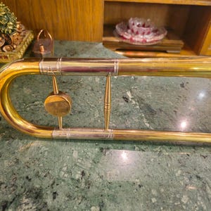 May include: A gold-colored trombone with a silver-colored slide and decorative accents. The trombone is lying on a green marble surface.