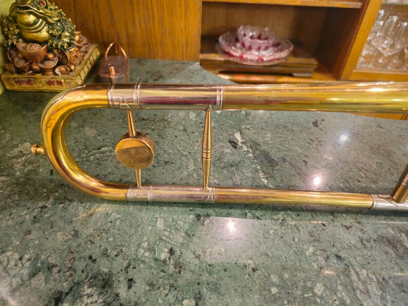 May include: A gold-colored trombone with a silver-colored slide and decorative accents. The trombone is lying on a green marble surface.