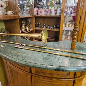 May include: A gold trombone with a bell-shaped mouthpiece rests on a green marble countertop. The trombone is in focus, while the background is blurred.