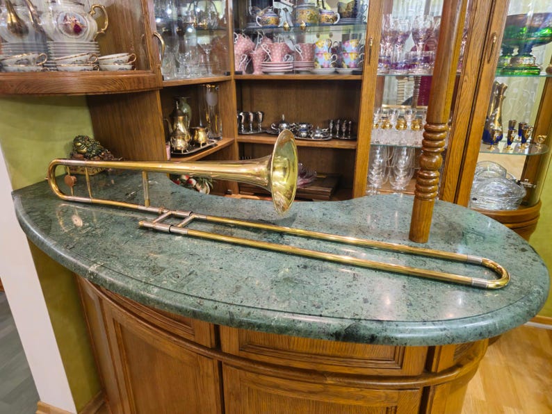 May include: A gold trombone with a bell-shaped mouthpiece rests on a green marble countertop. The trombone is in focus, while the background is blurred.