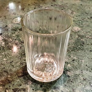 May include: A clear glass shot glass with a ribbed texture. The glass is empty and sitting on a green and gray countertop.