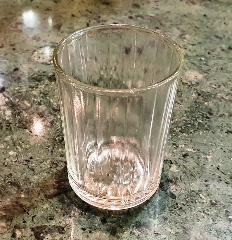May include: A clear glass shot glass with a ribbed texture. The glass is empty and sitting on a green and gray countertop.
