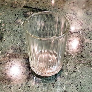 May include: A clear glass shot glass with a ribbed design. The glass is empty and sitting on a green and white marble surface.