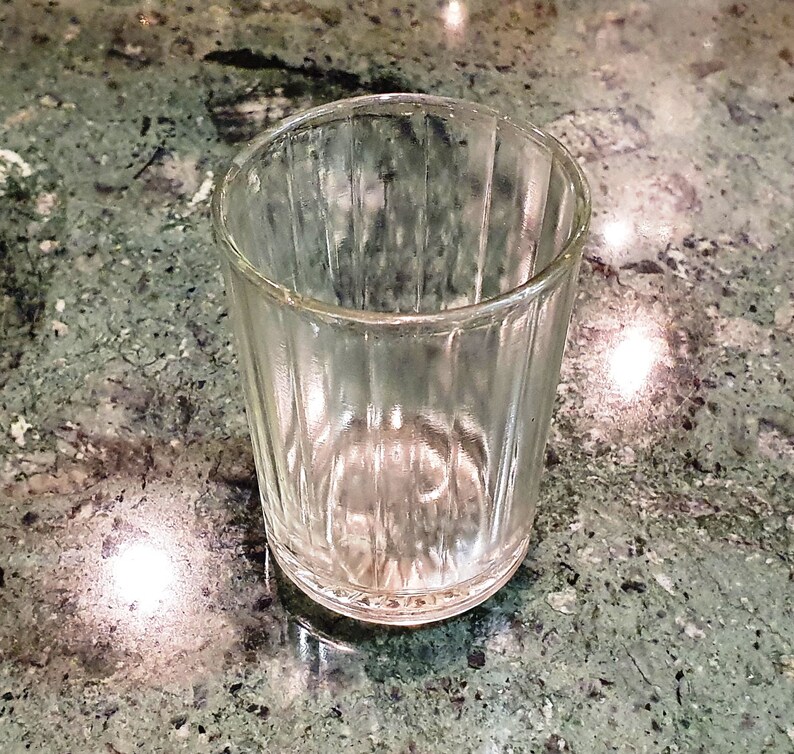 May include: A clear glass shot glass with a ribbed design. The glass is empty and sitting on a green and white marble surface.
