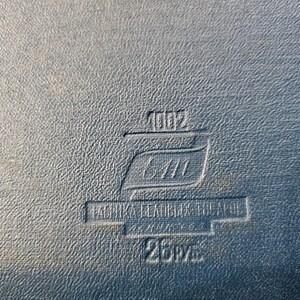 May include: A close-up of a black leather-bound book with a embossed label that reads "1002 6411 FABRIKA  BLANKA  CEAPOL 25PV", indicating it is a vintage book.