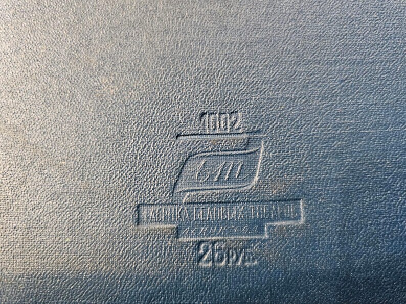 May include: A close-up of a black leather-bound book with a embossed label that reads "1002 6411 FABRIKA  BLANKA  CEAPOL 25PV", indicating it is a vintage book.