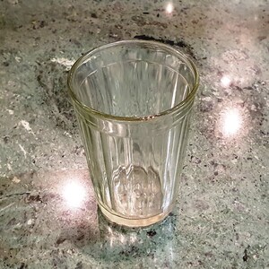 May include: A clear glass with vertical ridges, commonly known as a "shot glass" or "faceted glass", sits on a green and gray speckled surface.