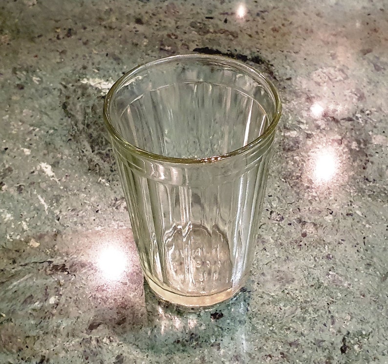 May include: A clear glass with vertical ridges, commonly known as a "shot glass" or "faceted glass", sits on a green and gray speckled surface.