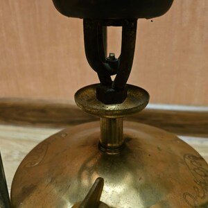 K&ouml;nnte beinhalten: Ein Vintage-Campingkocher aus Messing mit einem schwarzen Metallbrenner und einem schwarzen Metallst&auml;nder. Der Kocher hat einen runden Sockel und einen erh&ouml;hten Brenner.